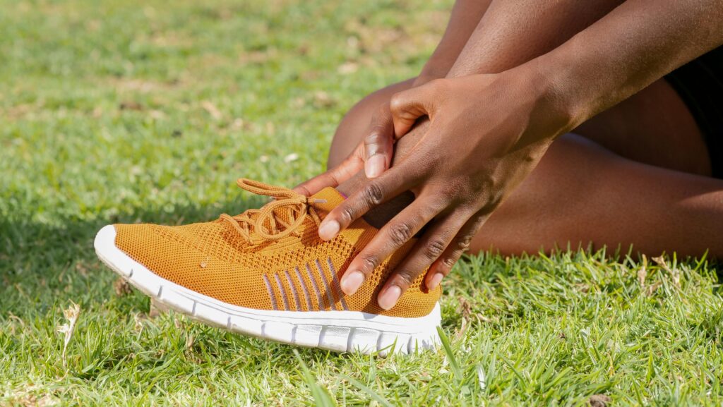 Black foot in yellow running shoe, hand holding ankle in apparent pain. Grass background.