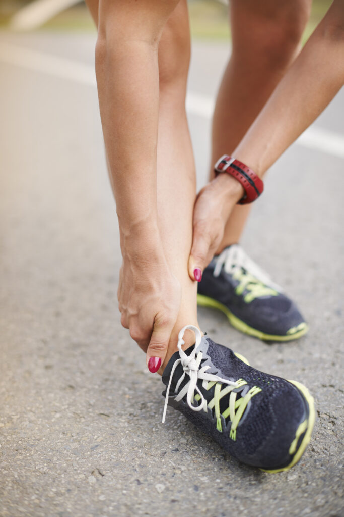 A close-up image of a woman experiencing ankle pain. She is wearing running shoes and is grasping her ankle with both hands.
